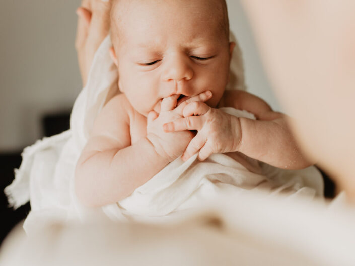 Ein Baby, was seine eigene Finger in den Mund steckt und auf dem Arm seiner Mama ist